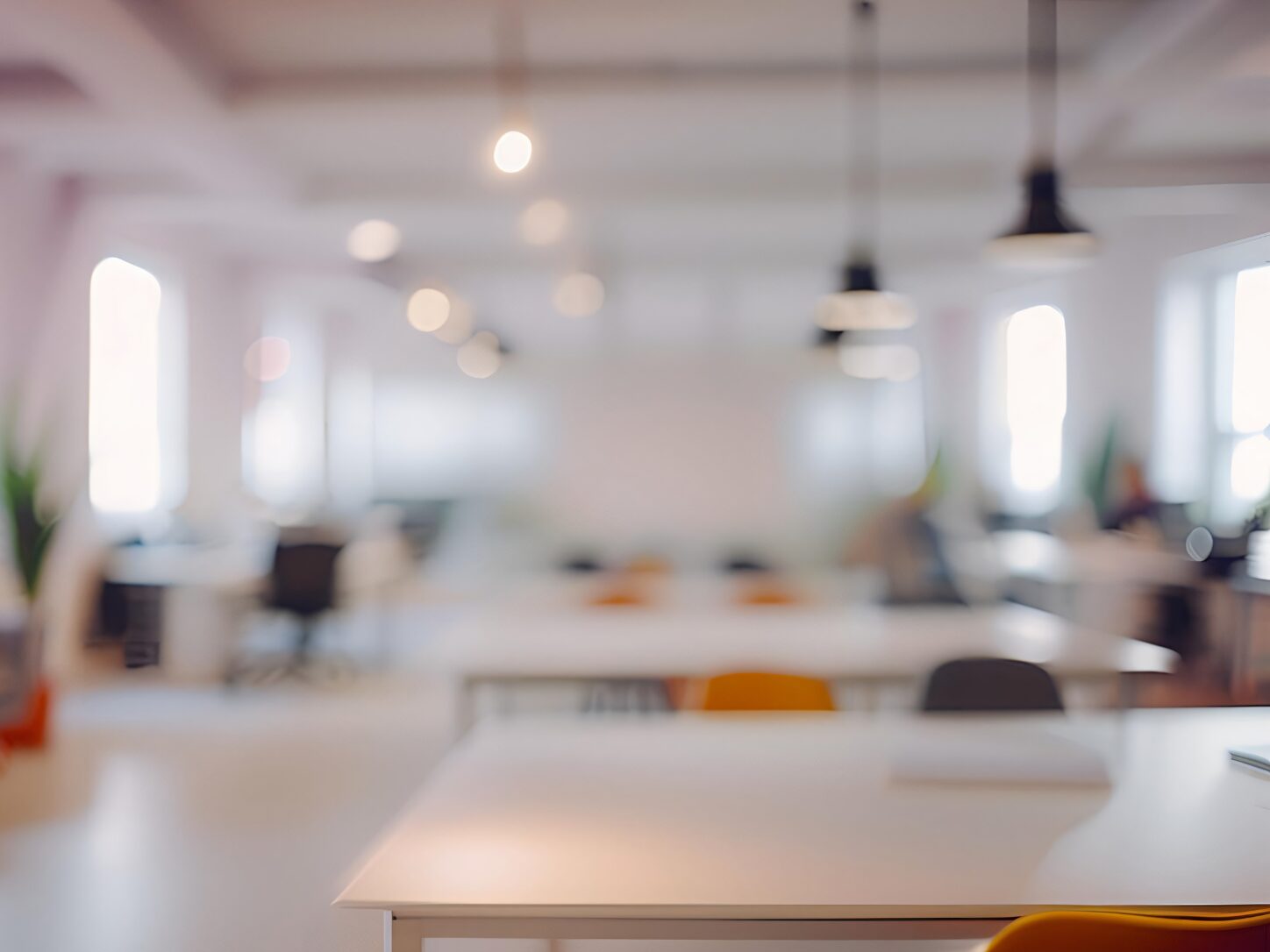 Soft-focus view of a bright open-plan office with desks, chairs and pendant lighting. Large windows fill the workspace with natural light, creating a clean, modern and spacious office environment.