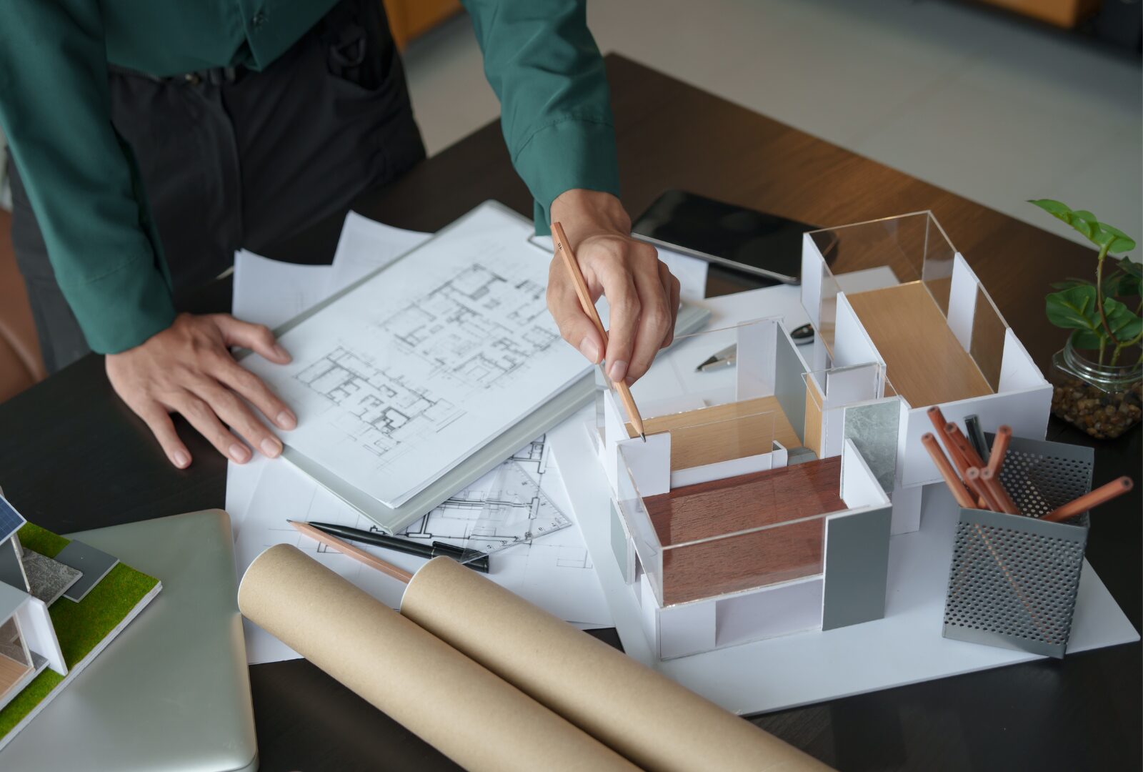 Architect reviewing architectural drawings and a scale model of a building, using a pencil to point out design details. Floor plans, rolled blueprints and design tools are spread across the desk, illustrating the planning and development of a building project.
