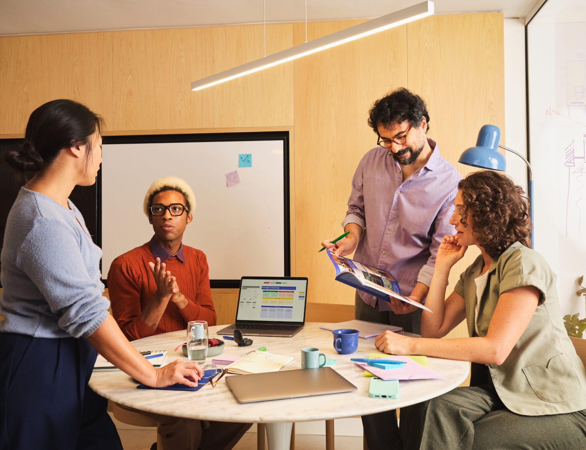 Diverse team of professionals collaborating around a meeting table in a modern office, discussing ideas and reviewing documents. A laptop, notebooks, sticky notes and presentation materials are spread across the table, highlighting teamwork and collaborative workspace design.